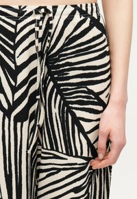 Κοντινό πλάνο σε σορτς με μοτίβο φύλλων σε ασπρόμαυρο, με ρυθμιζόμενο κορδόνι στη μέση, που δείχνει το δεξί χέρι ενός ατόμου να ακουμπάει δίπλα στο ρούχο.