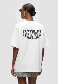 T-shirt bianca oversized in cotone con una grafica nera "BERLINO" sulla schiena. Il modello indossa occhiali da sole e una mini gonna texturizzata.