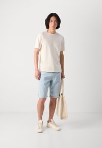 Cremefarbenes Baumwoll-T-Shirt mit kurzen Ärmeln und blauem Logo, hellblaue Denim-Shorts, beiger Einkaufstasche und beigen Sneakers, alles im lässigen Stil.