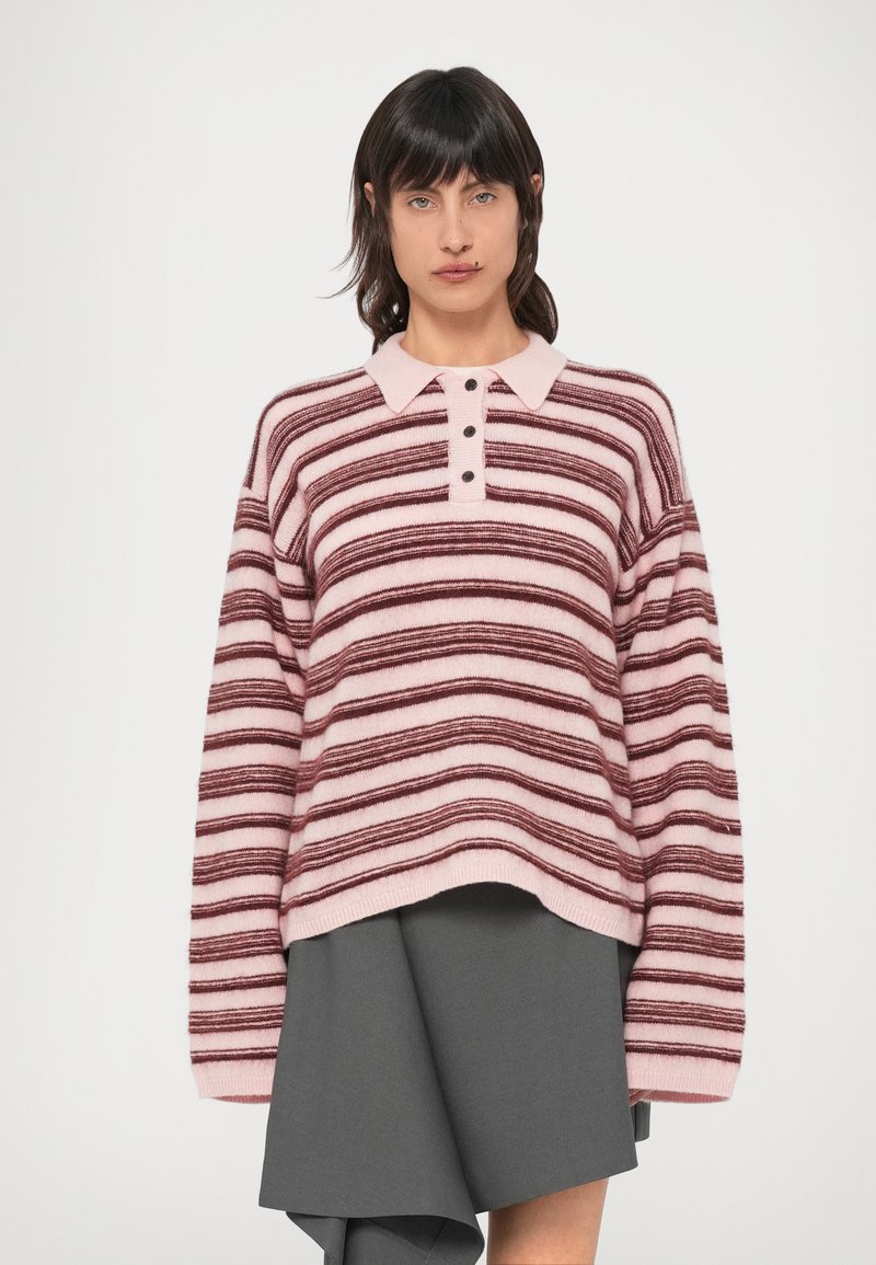 Ριγέ πουλόβερ σε ροζ και μπορντό, με γιακά, πλακέτα με τέσσερα κουμπιά και φαρδιά εφαρμογή. Συνδυάζεται με φούστα σε γκρι αποχρώσεις με στρώσεις.