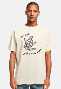 Cremefarbener Baumwoll-T-Shirt mit einem schwarzen Grafik eines Hundes auf einem Surfbrett und dem Text "Wir sehen uns auf der nächsten Welle." Kurze Ärmel, lässige Passform.