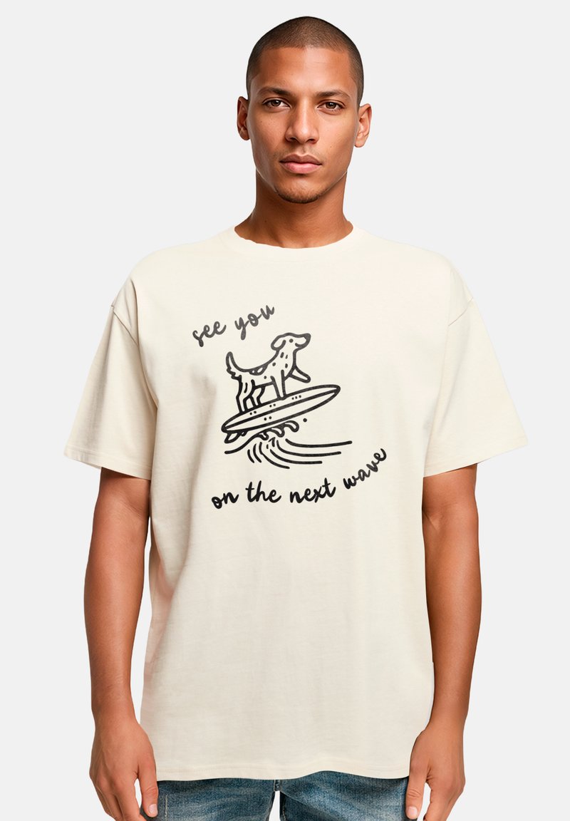 Cremefarbener Baumwoll-T-Shirt mit einem schwarzen Grafik eines Hundes auf einem Surfbrett und dem Text "Wir sehen uns auf der nächsten Welle." Kurze Ärmel, lässige Passform.