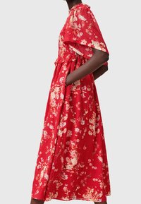 Femme portant une robe midi rouge à fleurs avec manches bouffantes courtes et corsage froncé, mains dans les poches latérales, vue de profil.