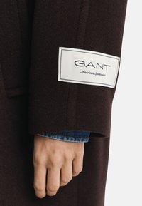 Bruine wollen jas met een wit GANT-label op de mouw, die een gladde textuur en op maat gesneden vorm toont, gedragen over een denimbloes.