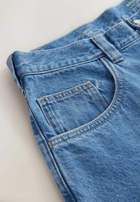Gros plan sur un jean en denim bleu montrant la poche avant, une passants de ceinture, et l'étiquette de taille visible "NEXT 34R" à la ceinture.