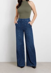 Olivgrön ribbad bodysuit som kombineras med höga vida mörkblå denimbyxor; har sidofickor och strukturerad tyg.