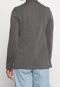 Blazer gris con un diseño estructurado, mangas largas y cuello con muescas. Tela suave y ajuste entallado, combinado con jeans de color azul claro.