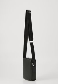 Lille sort rektangulær crossbody-taske med justerbar stofskulderrem og sølvfarvet metalhardware på en hvid baggrund.