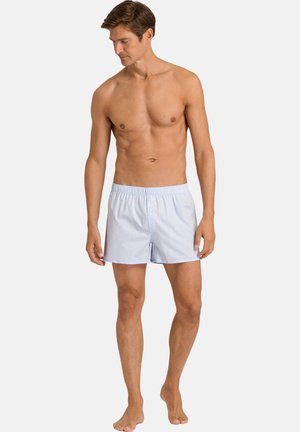 Mand stående barfodet iført lyseblå boksershorts med elastisk linning, kigger ned mod sin venstre side mod en ensfarvet baggrund.