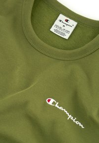T-shirt en coton olive avec col rond, arborant un logo "Champion" brodé en blanc et une étiquette d'entretien à l'intérieur du col.