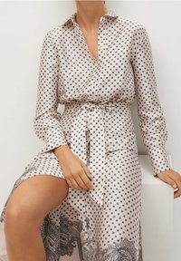 Donna seduta che indossa un abito lungo a maniche, di colore beige, con pois neri e motivi floreali, legato in vita con una cintura coordinata.