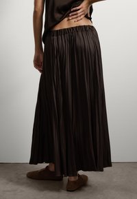 Bruine geplooide rok met een elastische tailleband, reikt tot halverwege de kuit. In combinatie met bruine instapschoenen. Eenvoudig, vloeiend ontwerp.