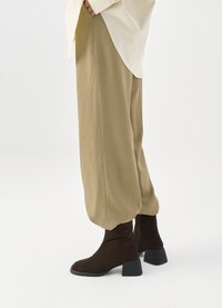 Brown suede ankle boots with a chunky black heel, paired with loose, light beige relaxed-fit trousers.  White top visible at the waist.