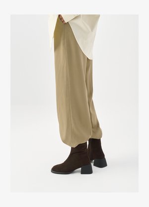 Bottines en daim marron à talon noir épais, accompagnées d'un pantalon beige clair à coupe décontractée. Un haut blanc est visible au niveau de la taille.