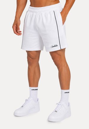 Uomo che indossa pantaloncini sportivi bianchi con piping nero, calzini bianchi e scarpe da ginnastica bianche, in piedi con le mani in tasca.
