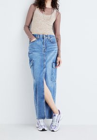 Denim maxi skirt with side slit, two front pockets, and top button closure, paired with a beige tank top and purple-accented sneakers.