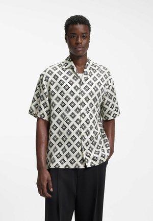 Man wearing short-sleeve, button-up shirt with black floral diamond pattern and black trousers, standing with one hand in pocket.