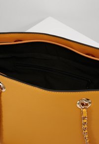 Żółta torebka o teksturowanej powierzchni, czarnym wnętrzu i złotym łańcuszku jako ozdobie. Wewnątrz znajduje się kieszeń zapinana na zamek do przechowywania.