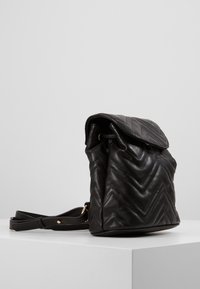 Schwarzer, gesteppten Leder-Rucksack mit Klappenverschluss, der ein Zickzackmuster aufweist und einen verstellbaren Riemen hat. Akzente in goldfarbener Hardware.