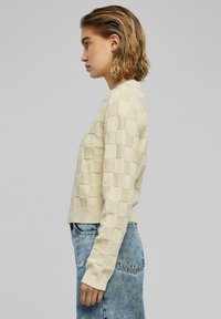Beige gebreide trui met een textuur van ruitjespatroon, voorzien van geribde mouwen en zoom, gedragen met hoogwaisted, distressed denim jeans.