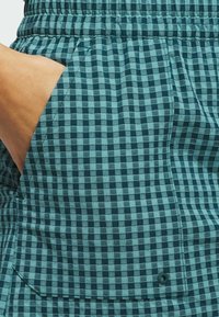 Turkusowo-granatowa tkanina w kratę z kieszenią, elastycznym paskiem w talii i widocznymi szwami. Tekstura wydaje się gładka i lekka.