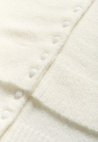 Fehér, textúrált kötött kardigán, amely puha, bolyhos anyagból készült. Különleges részletei közé tartoznak az egyenletesen elhelyezett, szövettel borított gombok a szélén.