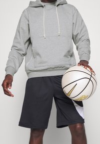 Personne portant un sweat à capuche gris et un short noir tenant un ballon de basket Nike blanc et or près de la hanche.