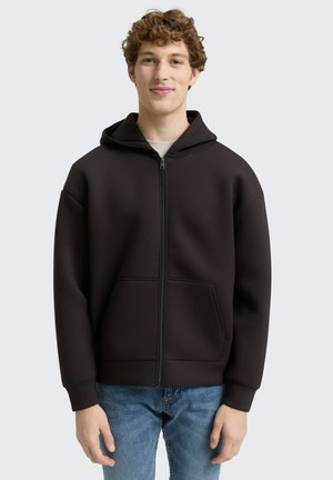 Schwarze Kapuzenjacke mit Reißverschluss aus strukturiertem Stoff, mit einer Kapuze, zwei Fronttaschen und gerippten Bündchen. Getragen über einem hellen Hemd mit blauer Jeans.