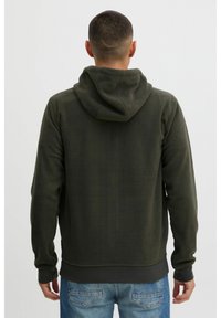 Sudadera de felpa verde oscuro con capucha, mangas largas y bajo acanalado. Presenta una textura suave y un ajuste relajado, combinada con vaqueros azules.