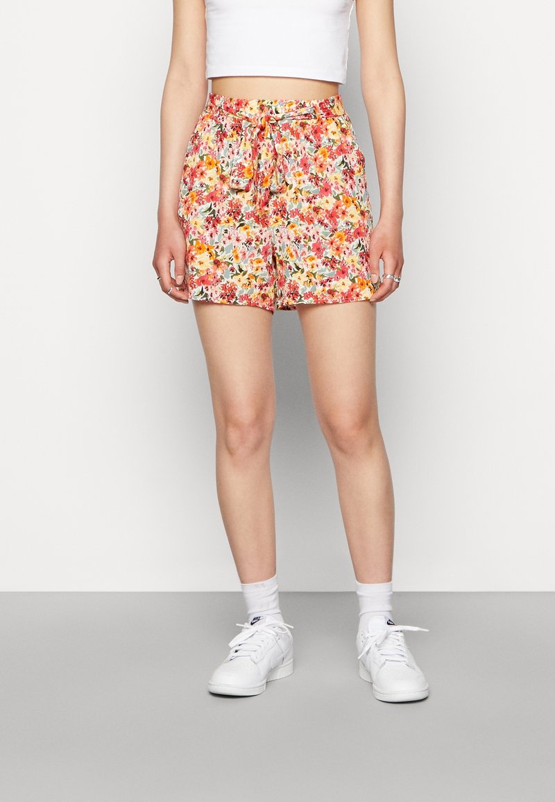 Femme portant un short taille haute à motifs floraux avec une ceinture nouée à la taille, associée à des baskets blanches, des chaussettes blanches et un haut court blanc.