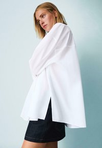 Camicia bianca oversize con maniche lunghe, caratterizzata da spacchi laterali e una vestibilità comoda, abbinata a pantaloni corti in denim scuro, su uno sfondo chiaro.