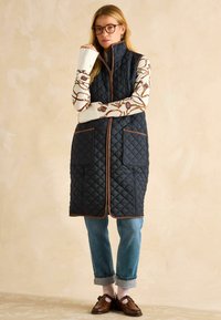 Izšūta jahtu veste ar augstu apkakli, brūnu apmalīti un divām priekšējām kabatām. Apvienota ar rakstainu džemperi, rullētiem džinsiem un brūnām kurpēm.