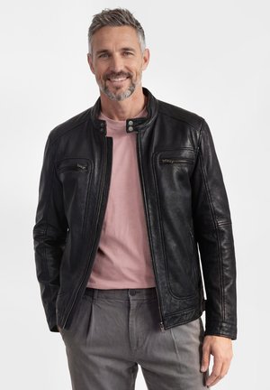 Lachende man met grijs haar, gekleed in een zwarte leren jas over een roze shirt en grijze broek, tegen een effen witte achtergrond.