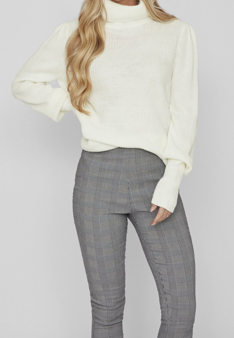 Femme portant un pull en maille à col roulé blanc et un pantalon ajusté à carreaux gris taille haute, avec de longs cheveux blonds tombant sur une épaule.