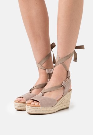 Jalat yang mengenakan sandal wedge espadrille suede krem dengan tali yang melilit di pergelangan kaki diikat menjadi busur di latar belakang putih.