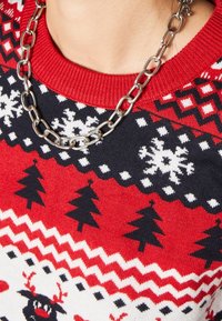 Pull rouge et marine avec des motifs festifs incluant des arbres, des flocons de neige et des rennes, accompagné d'un collier en chaîne argentée.