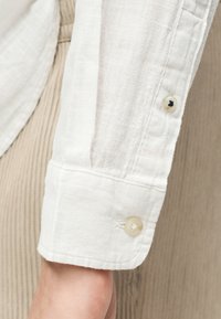 Μανίκι λευκού πουκαμίσου από λινό με μανσέτα με κουμπί, εμφανιζόμενο σε καρπό, πάνω σε φόντο από μπεζ ριγωτό ύφασμα.