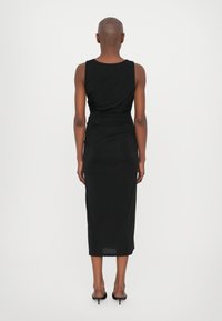 Robe midi noire sans manches avec un design drapé et une texture lisse. Présente une taille cintrée et arrive au mollet, associée à des talons noirs.