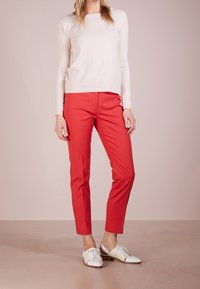 Jasnoróżowy sweter z długim rękawem w połączeniu z czerwonymi dopasowanymi spodniami. Model ma na sobie białe buty sznurowane. Minimalistyczny design z gładkimi teksturami.