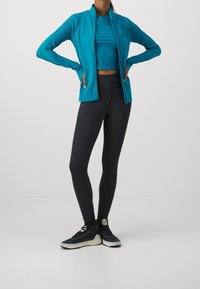 Giacca teal con zip, top fitted teal, leggins neri e sneakers nere. La giacca ha una texture liscia; i leggins sono elastici.