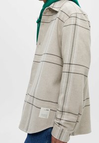 Beige shirt met ruitpatroon, gemaakt van textuurstof met witte en bruine lijnen. Voorzien van een groene capuchon en een label aan de onderkant.