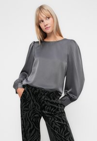 Grå satengbluse med puffede ærmer, rund hals og subtil glans. Sammen med sorte, mønstrede velourbukser med en tekstureret design.