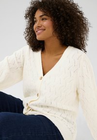 Cardigan lavorato a maglia crema con scollo a V, caratterizzato da un motivo a onda e tre bottoni sul davanti. Texture morbida, vestibilità rilassata e polsini a coste.