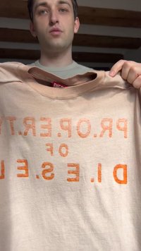 Tânăr ținând un hanorac bej deschis cu text decolorat portocaliu „P.R.O.P.E.R.T.Y OF D.I.E.S.E.L” în interior.