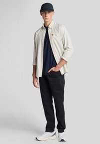 Camicia leggera button-up sopra una t-shirt blu navy, pantaloni neri e sneakers bianche. La camicia presenta un piccolo logo giallo.