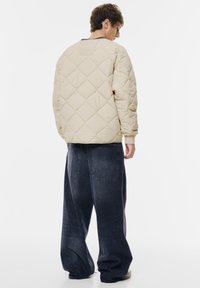 Veste matelassée beige avec un col rond et des poignets côtelés, associée à un jean bleu foncé à jambes larges avec des poches arrière.