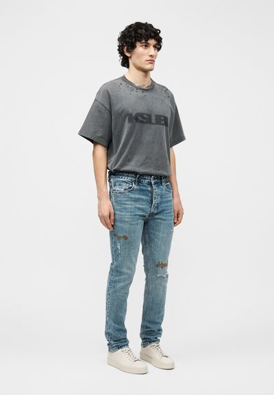 Νεαρός άνδρας όρθιος, φορώντας φθαρμένο, υπερμεγέθες γκρι T-shirt με τρύπες, ξεβαμμένα μπλε τζιν και λευκά αθλητικά παπούτσια, μπροστά σε μια απλή φόντο.