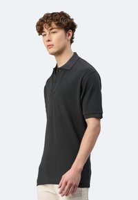Dunkelgraues Poloshirt mit einer Zweiknopf-Leiste, kurzen Ärmeln und einem strukturierten Stoff, getragen von einem männlichen Modell, das leicht zur Seite geneigt ist.