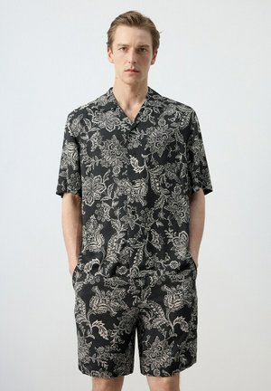 Hombre con camisa negra de manga corta y pantalones cortos a juego con estampado floral blanco, manos en los bolsillos, fondo neutro.