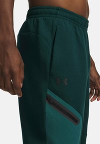 Grüne Sporthose aus weichem Material, mit einer Reißverschlusstasche, elastischem Bund und einem schwarzen Logo an der Seite.